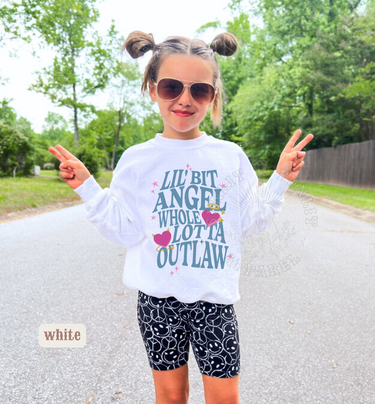 A young girl wearing sunglasses and a white sweatshirt with text printed on it, standing on a road with trees in the background.